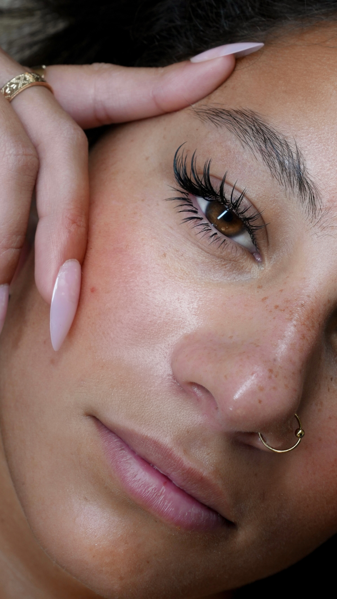 lose-up of a woman with long lash extensions, nude nails, freckles, and a gold nose ring, resting her hand against her face.