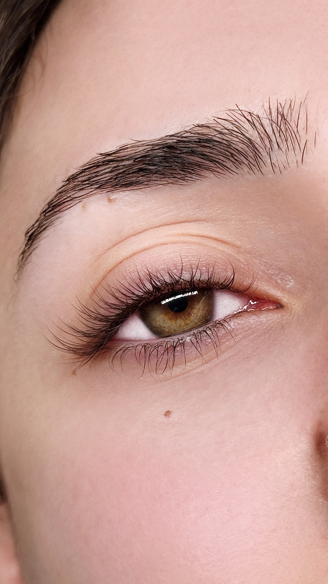Close-up of a person’s eye with long natural-looking eyelash extensions and a well-groomed brow. The lashes have a soft curl, and the person’s skin is smooth with a small beauty mark visible beneath the eye.
