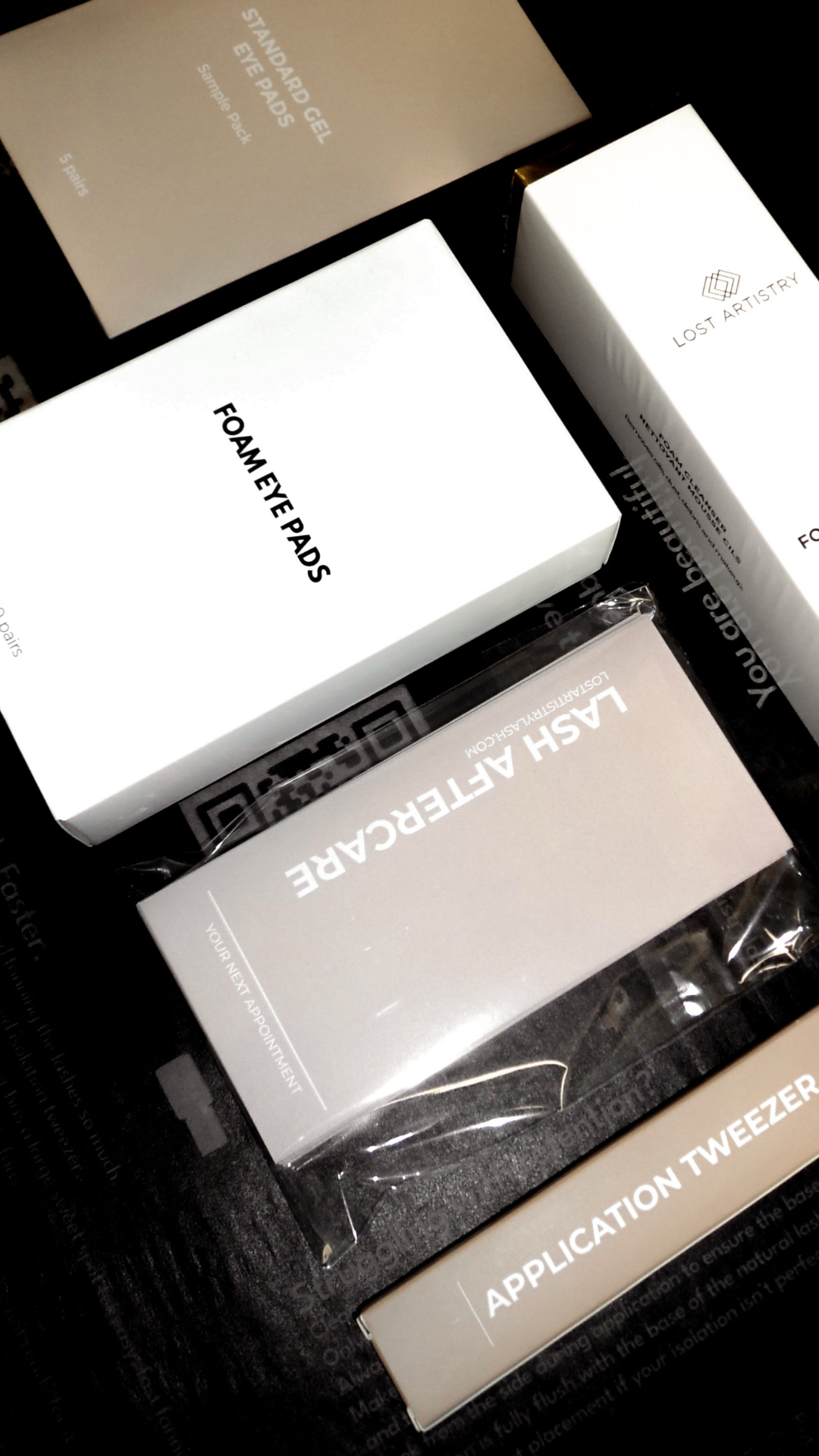 A flat lay of Lost Artistry Lash products displayed on a dark surface. The items include boxes labeled Foam Eye Pads, Lash Aftercare, Application Tweezer, and Shampoo Gel. The packaging is minimalist, with clean white boxes and light beige accents.
