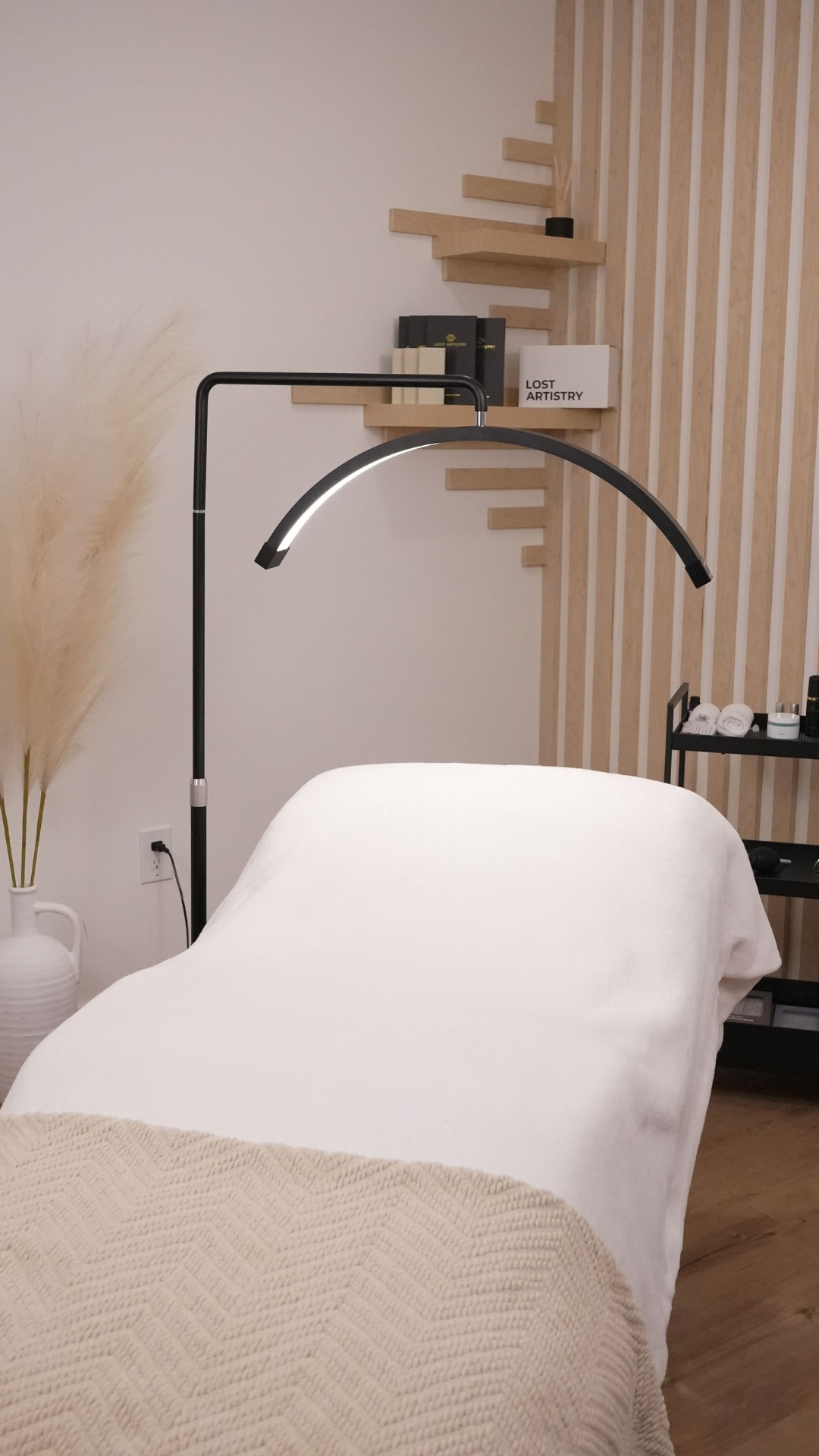 A clean lash studio setup with a treatment bed covered in white sheets and a beige knit blanket, lit by a curved LED lamp. Shelves with Lost Artistry products and décor are visible in the background, creating a modern and minimal aesthetic.