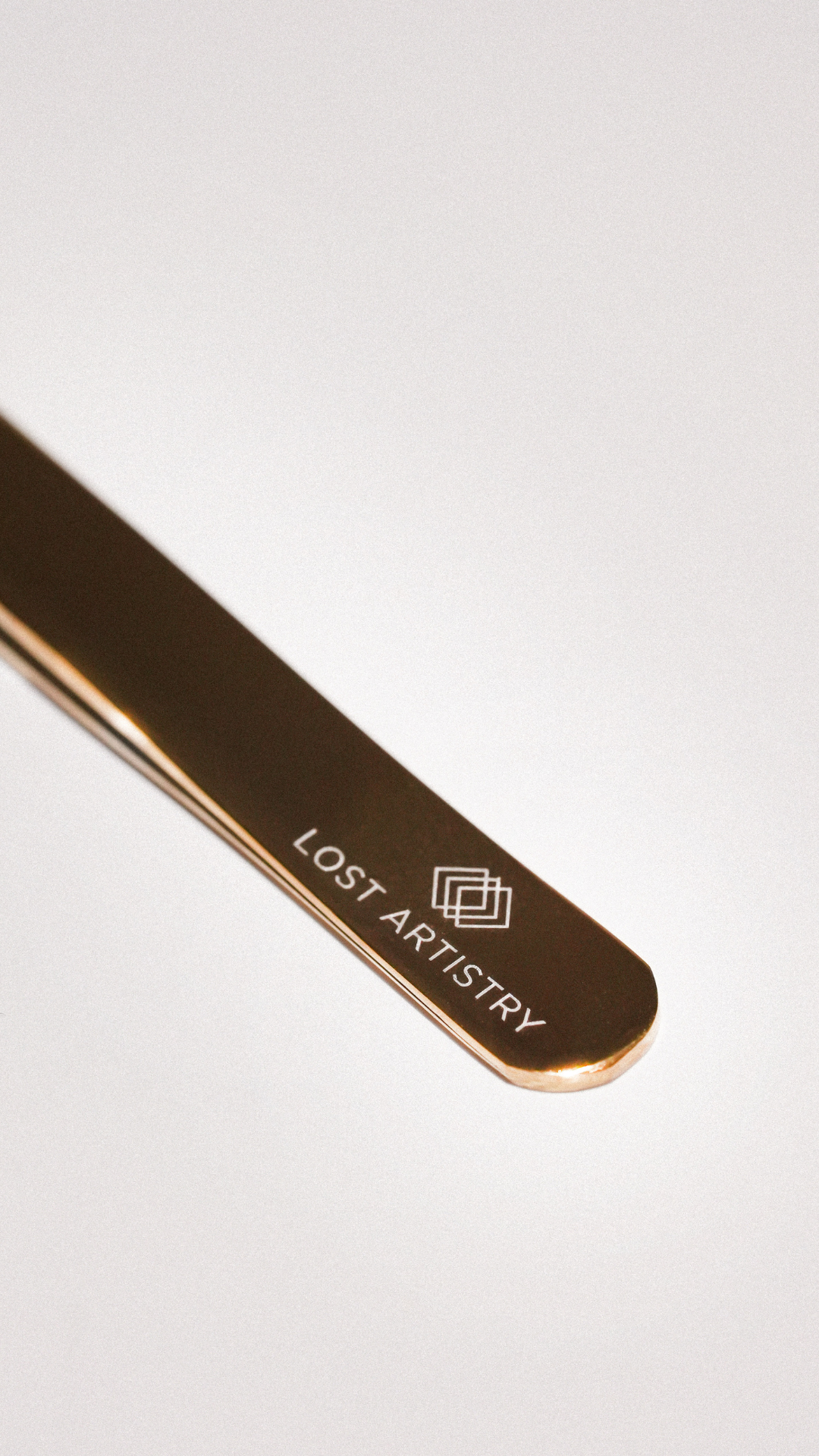 Close-up of a gold eyelash extension tweezer engraved with the Lost Artistry logo on a white background.