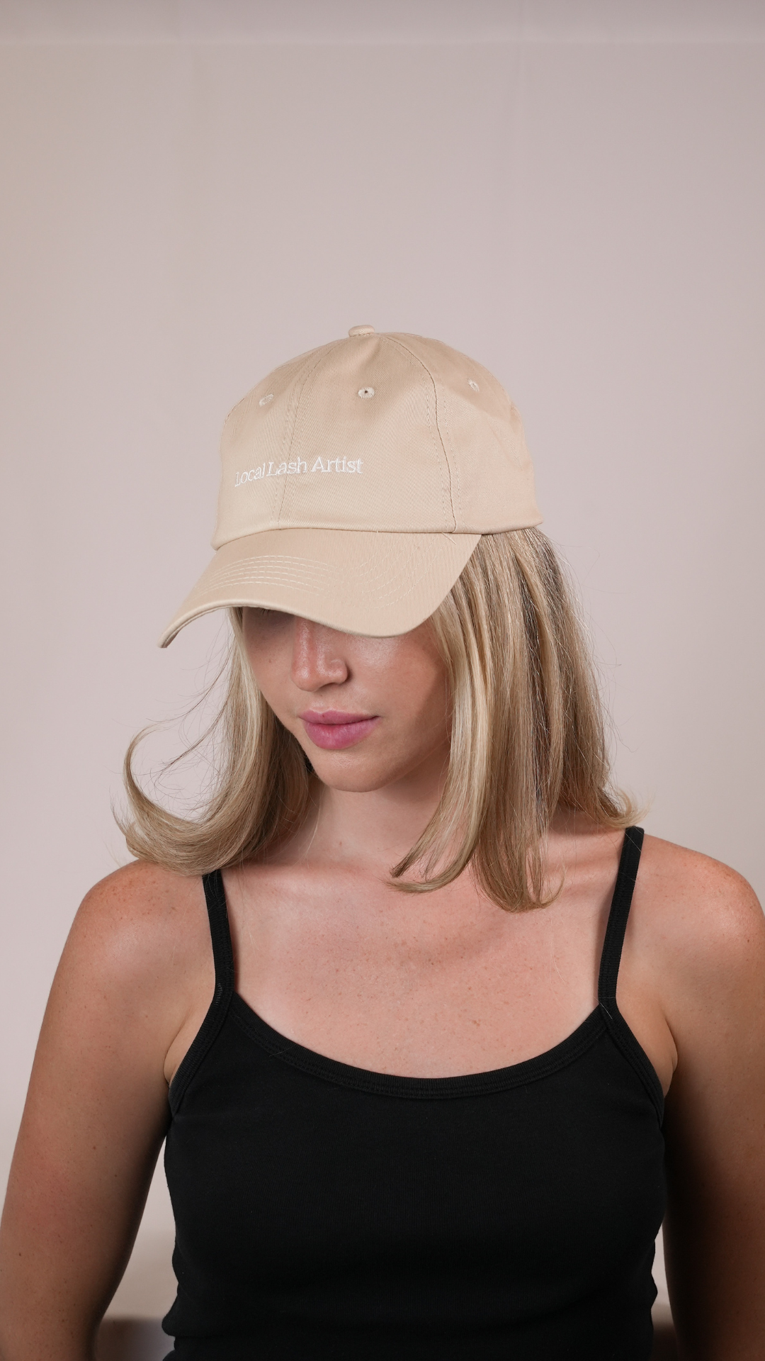 Woman wearing a beige baseball cap embroidered with the words “Local Lash Artist,” looking down slightly, with straight blonde hair, a black tank top, and a plain neutral background.