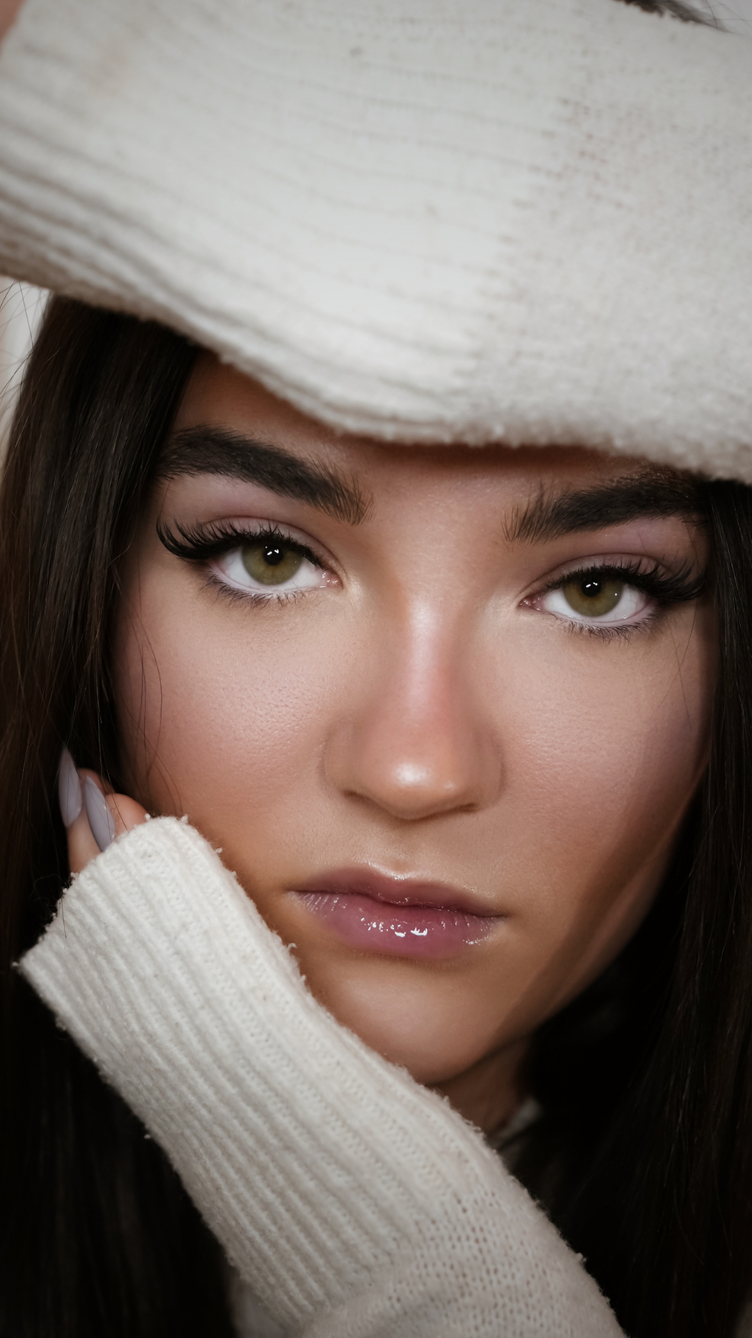 A close-up portrait of a woman with long dark hair and soft glam makeup, showcasing full eyelash extensions, wearing a cozy cream sweater with her hand resting near her face.