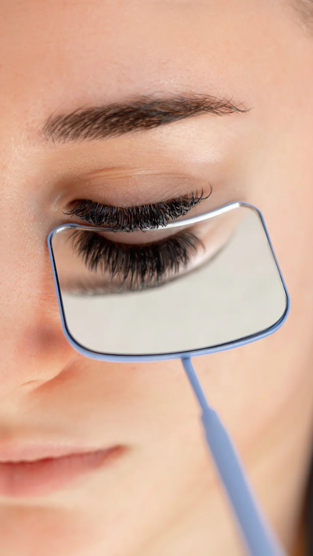 Close-up of a woman's eye with eyelash extensions reflected in a small handheld lash mirror during a lash application.