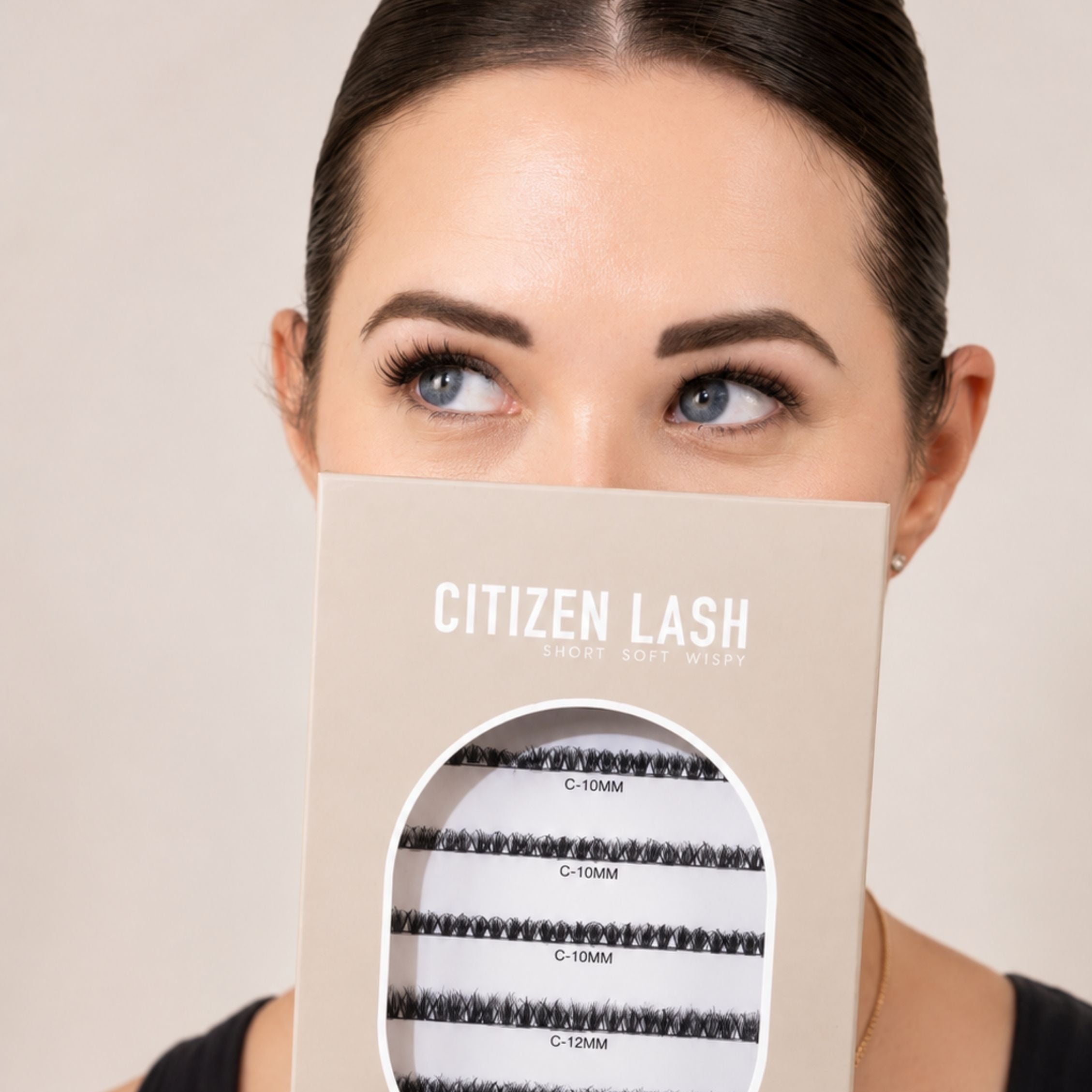 A woman holds a beige box of Citizen Lash false eyelashes in front of her face. The box features a clear oval window displaying black lash clusters labeled with different lengths (C-10MM, C-10MM, C-10MM, C-12MM).