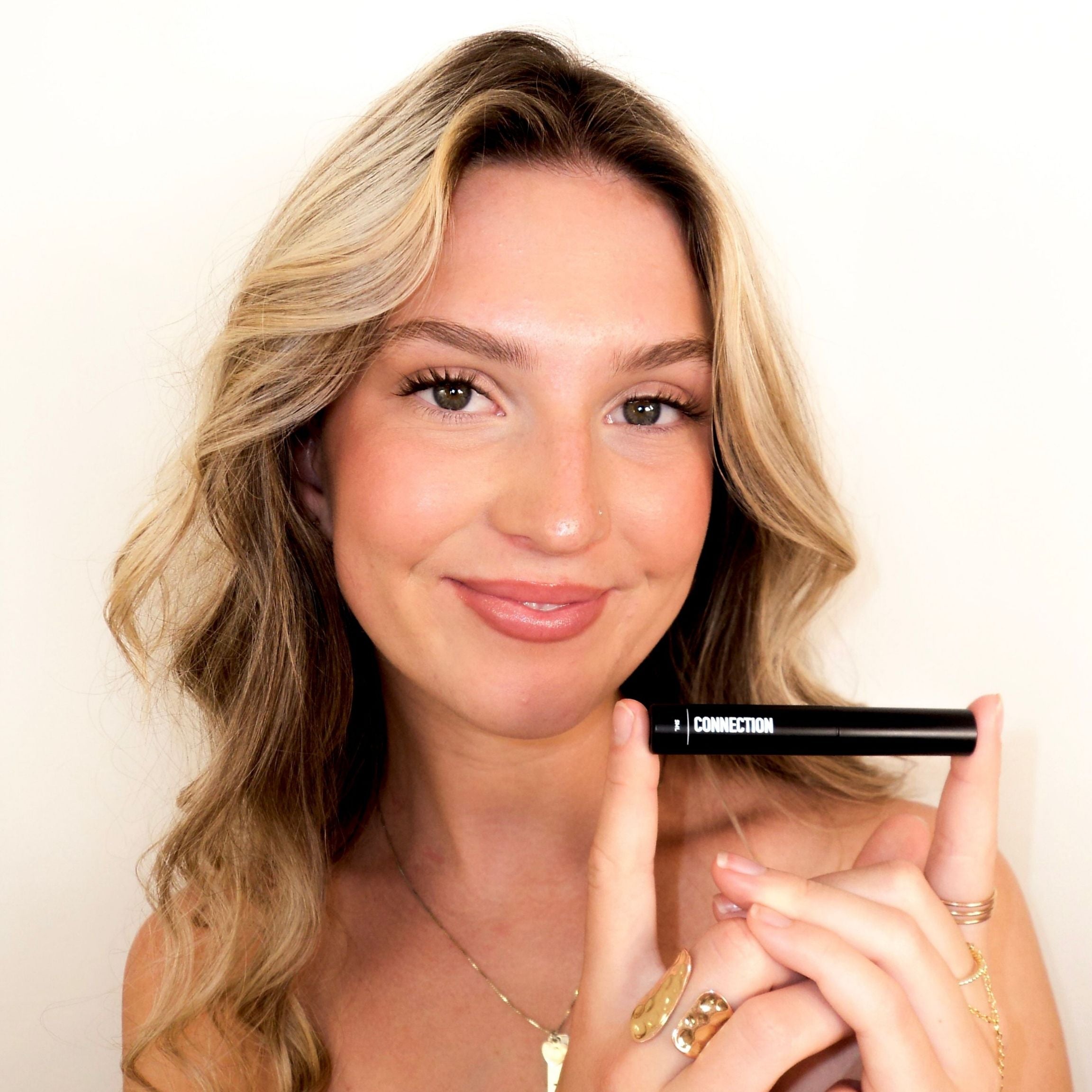 A woman with blonde hair holds a black eyeliner pencil with the brand name 'CONNECTION' visible, set against a plain white background.