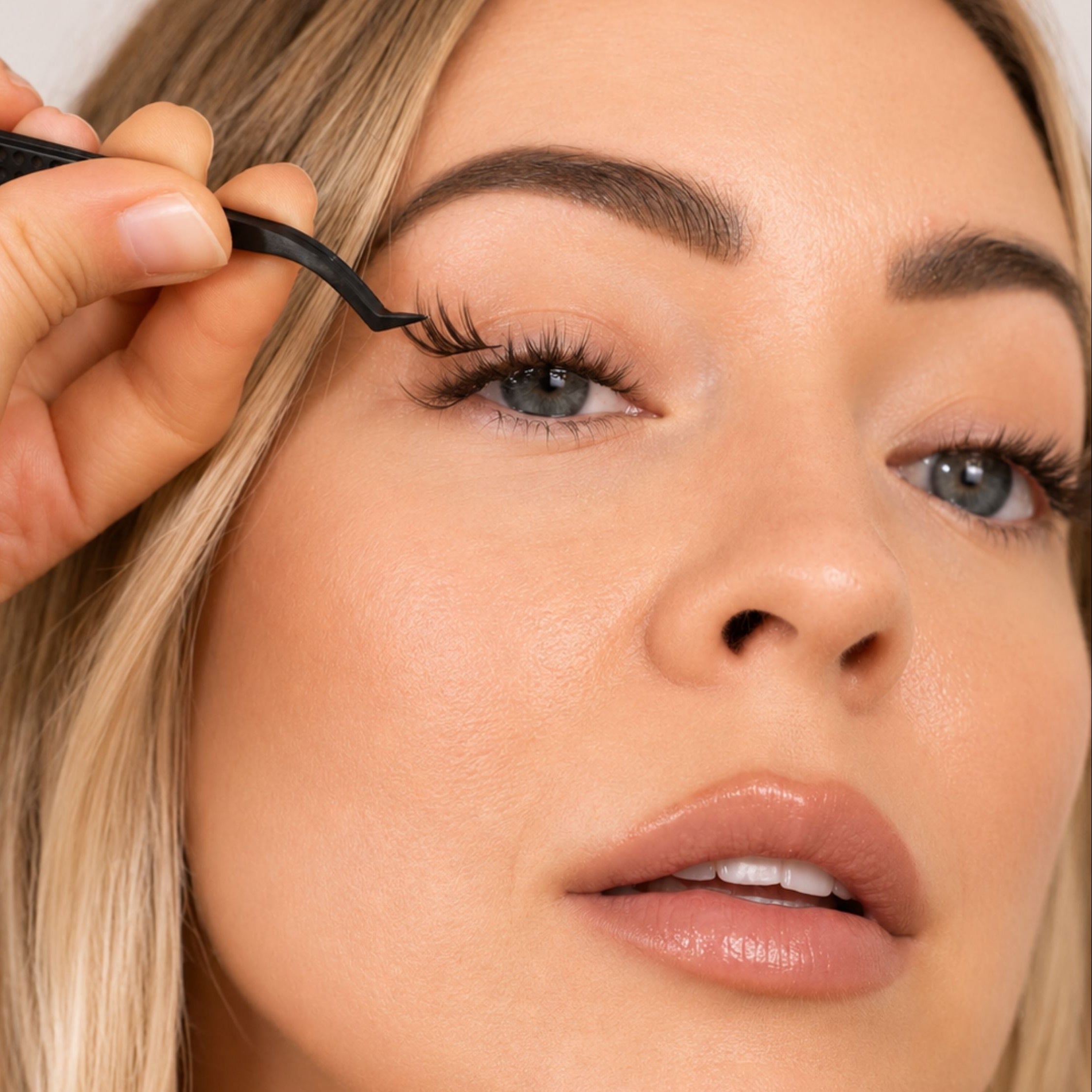 Close-up of a woman applying false eyelashes with a black tweezers tool, showing detailed application to her upper eyelashes.