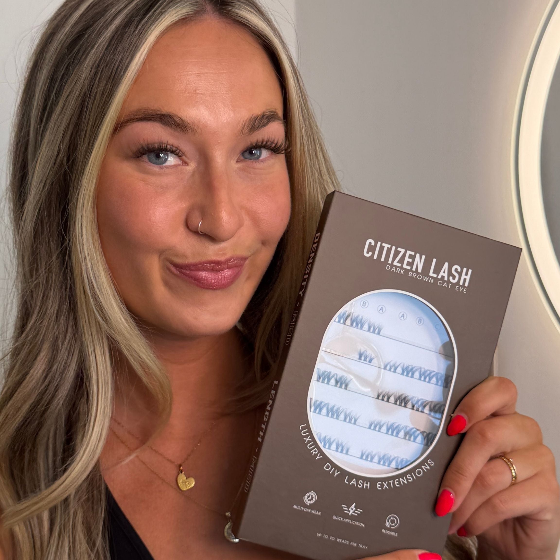 A woman holding a box of Citizen Lash Beauty Studio Cat Eye Luxury DIY Lash Extensions in front of a mirror with a ring light.