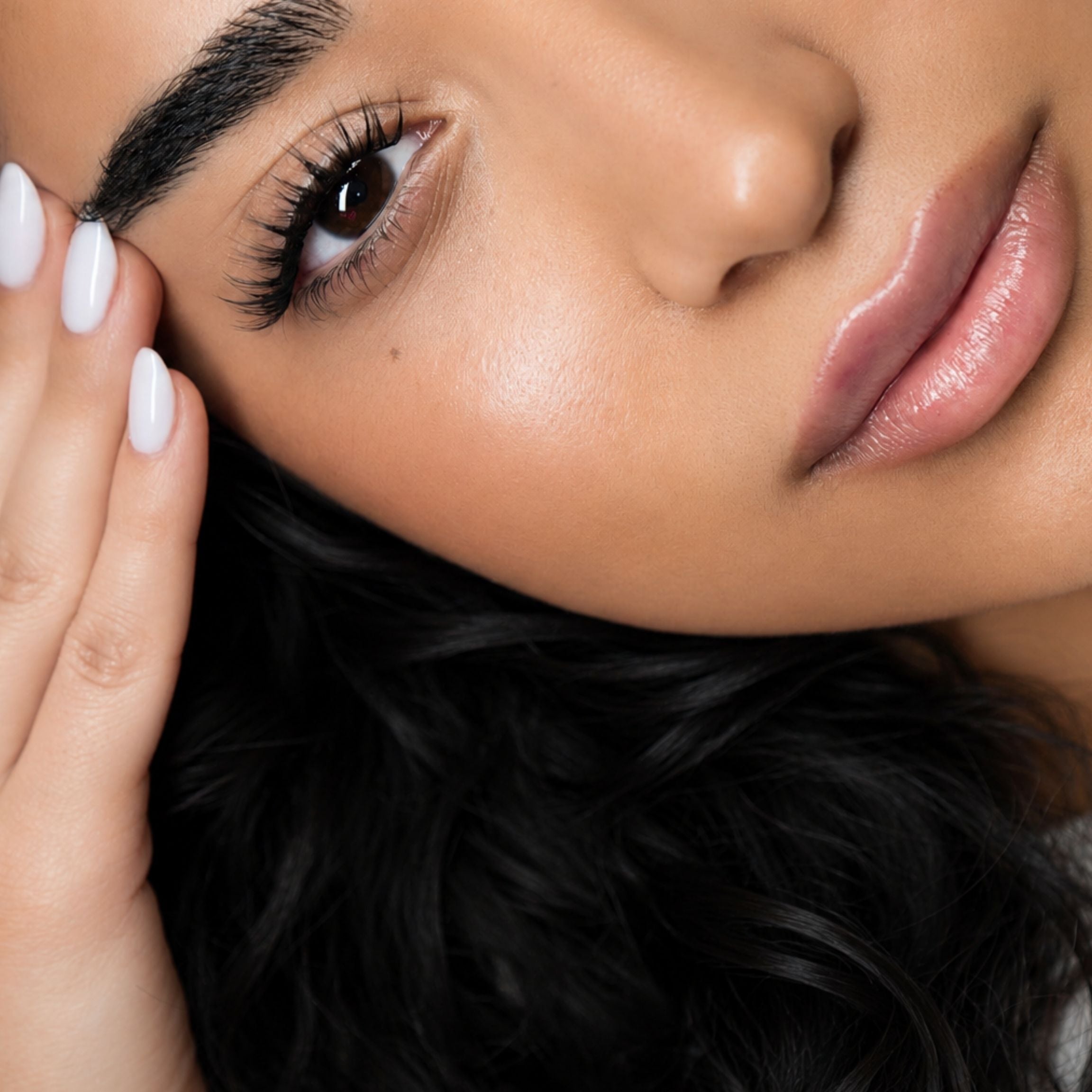 Close-up of a woman's face with long, glossy black eyelashes, defined eyebrows, and nude pink lips, with her hand resting near her temple.