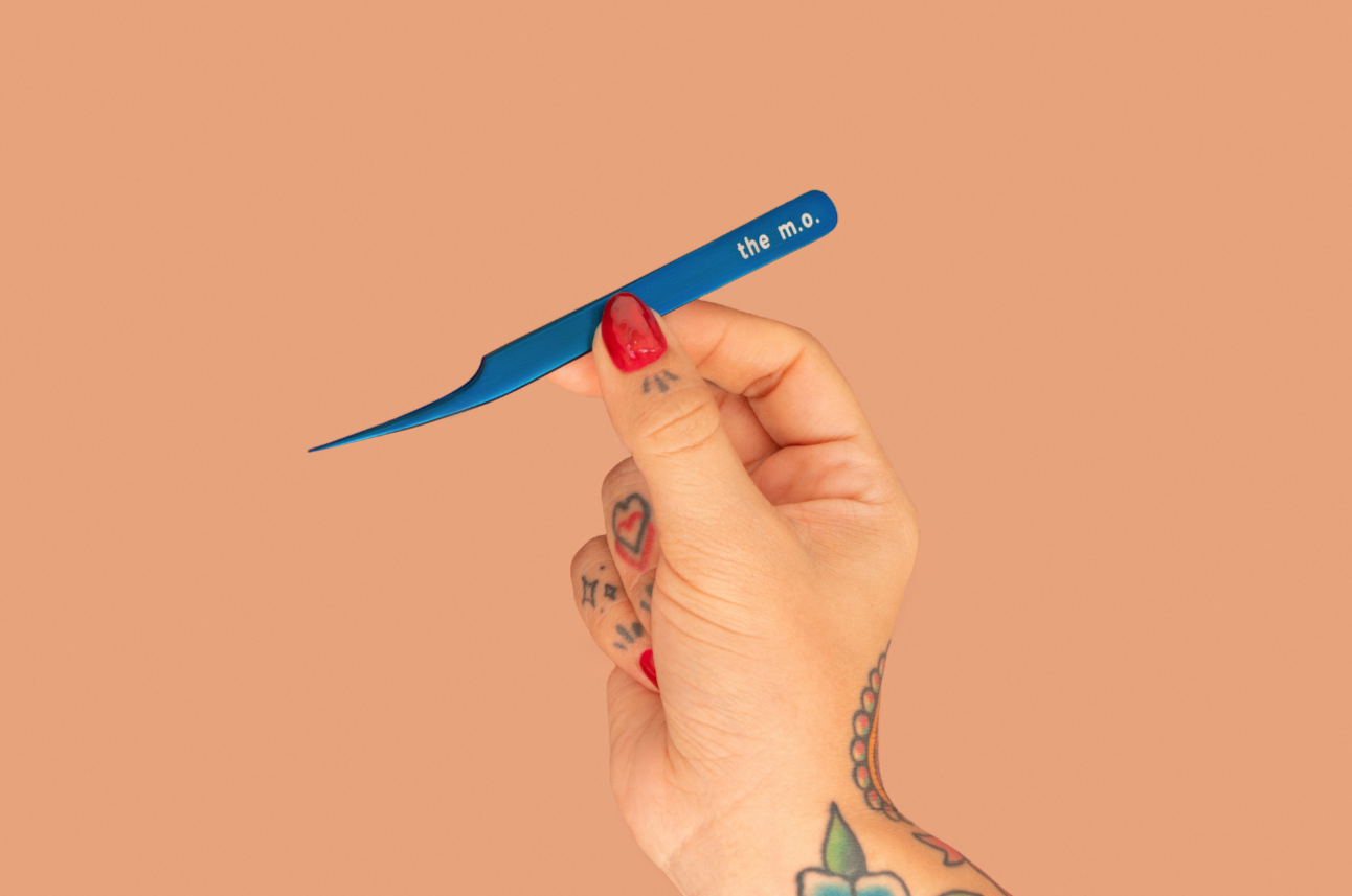 Hand holding a blue tweezer with 'the m.e.' branding against a beige background