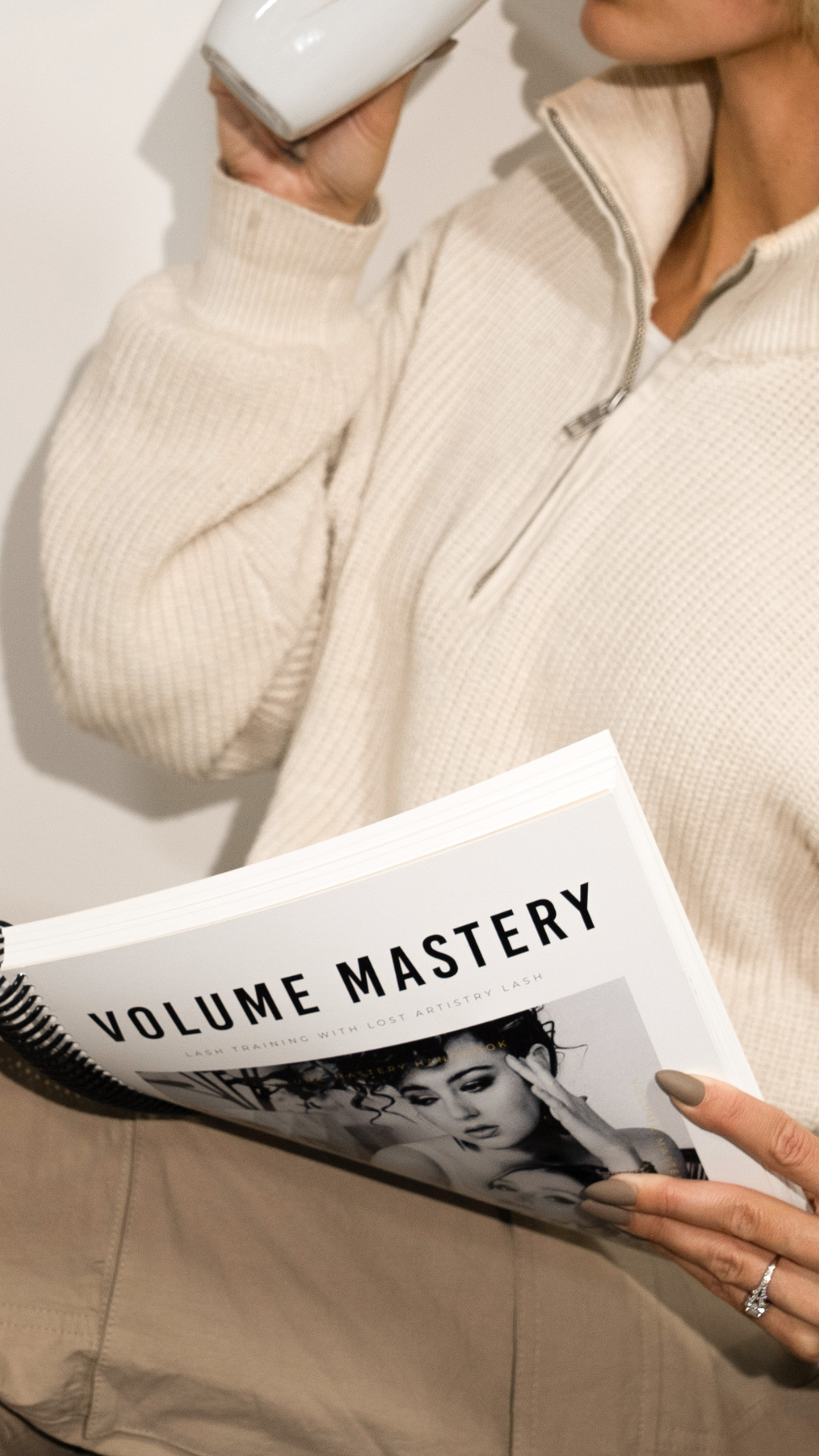 Person in a beige zip-up sweater holding a white mug and flipping through Lost Artistry’s Volume Mastery lash training manual.