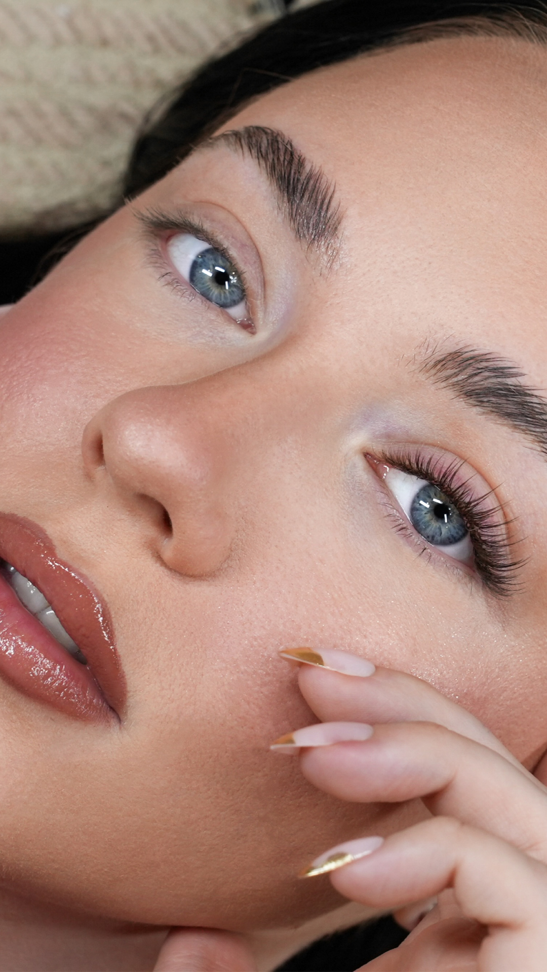 Close-up of a woman with blue eyes, glossy lips, and long natural-looking lash extensions, resting her hand near her face with manicured nails.