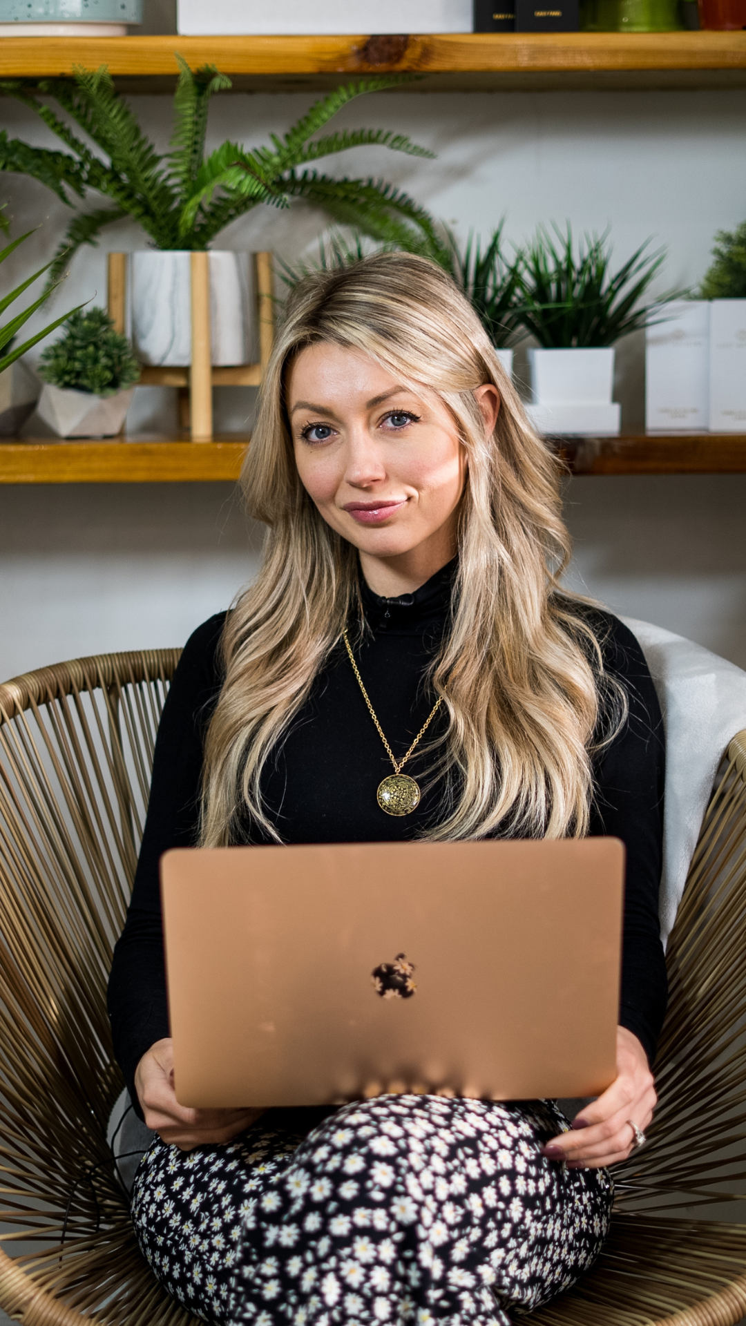 Smiling woman with long blonde hair sitting in a wicker chair, working on a rose gold laptop, with plants and shelves in the background.