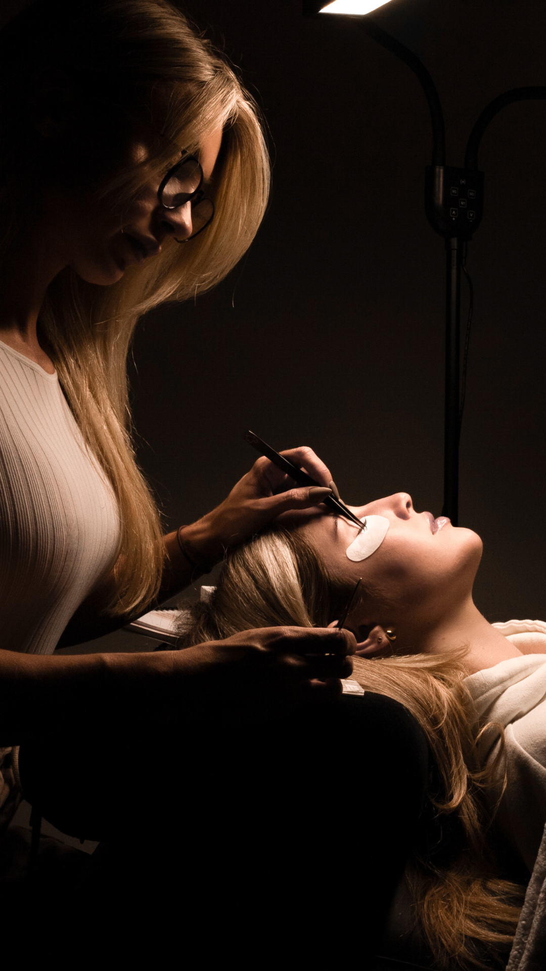 Lash artist applying eyelash extensions under professional salon lighting with eye pads for client comfort.