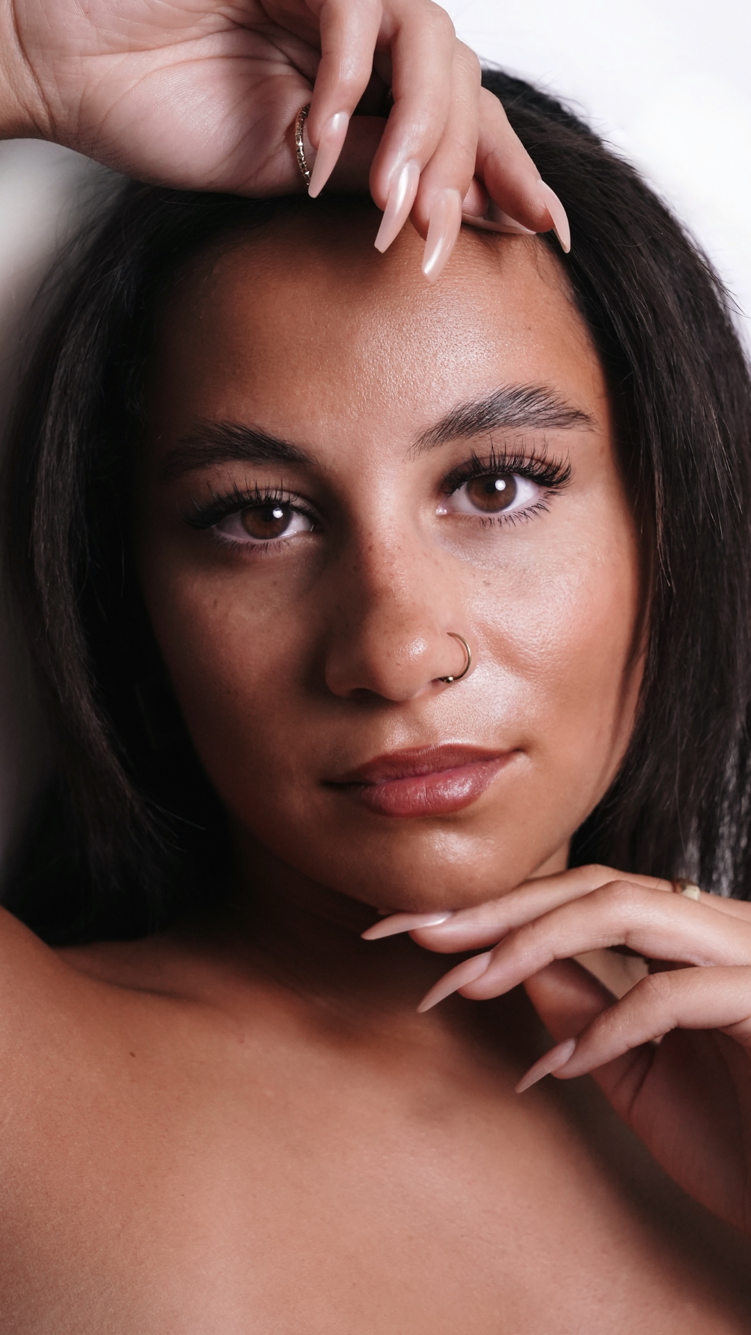 Close-up portrait of a woman with natural makeup, long lashes, and manicured nails gently resting on her face.