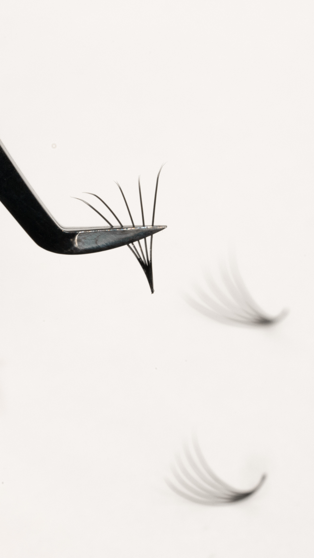 Close-up of metal tweezers holding a handmade eyelash fan against a white background, with additional lash fans blurred below.