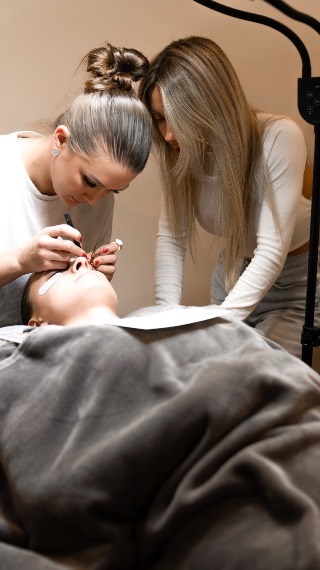 Lash artist applying extensions on a client lying under a blanket while another woman observes closely under a salon light