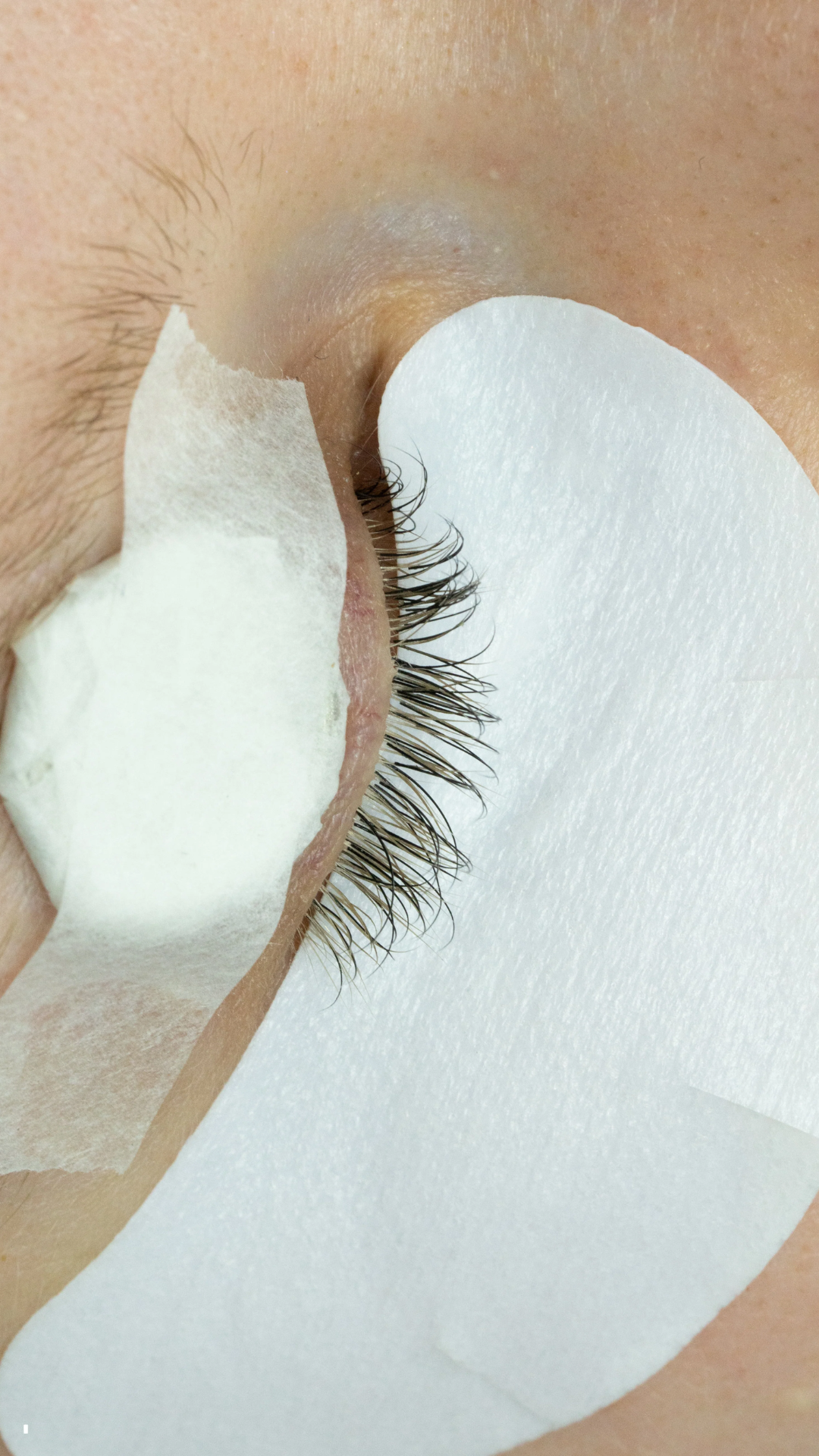 Close-up of an eye prepared for lash extensions, with under-eye pads and tape holding a coin in place to reduce fluttering