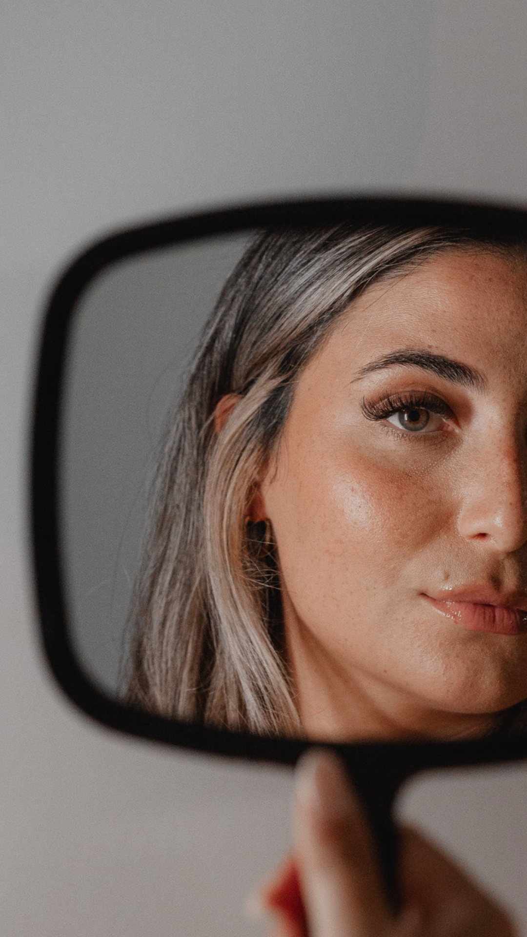 Close-up reflection of a woman’s face in a handheld mirror, showing natural makeup and long eyelashes