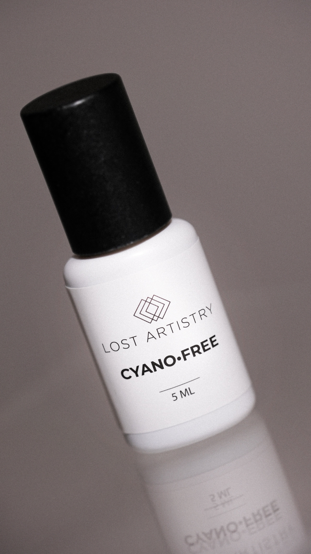 A small white 5 ml bottle of Lost Artistry’s Cyano-Free adhesive with a black cap, standing on a reflective surface. The label shows the brand logo and product name in black text.