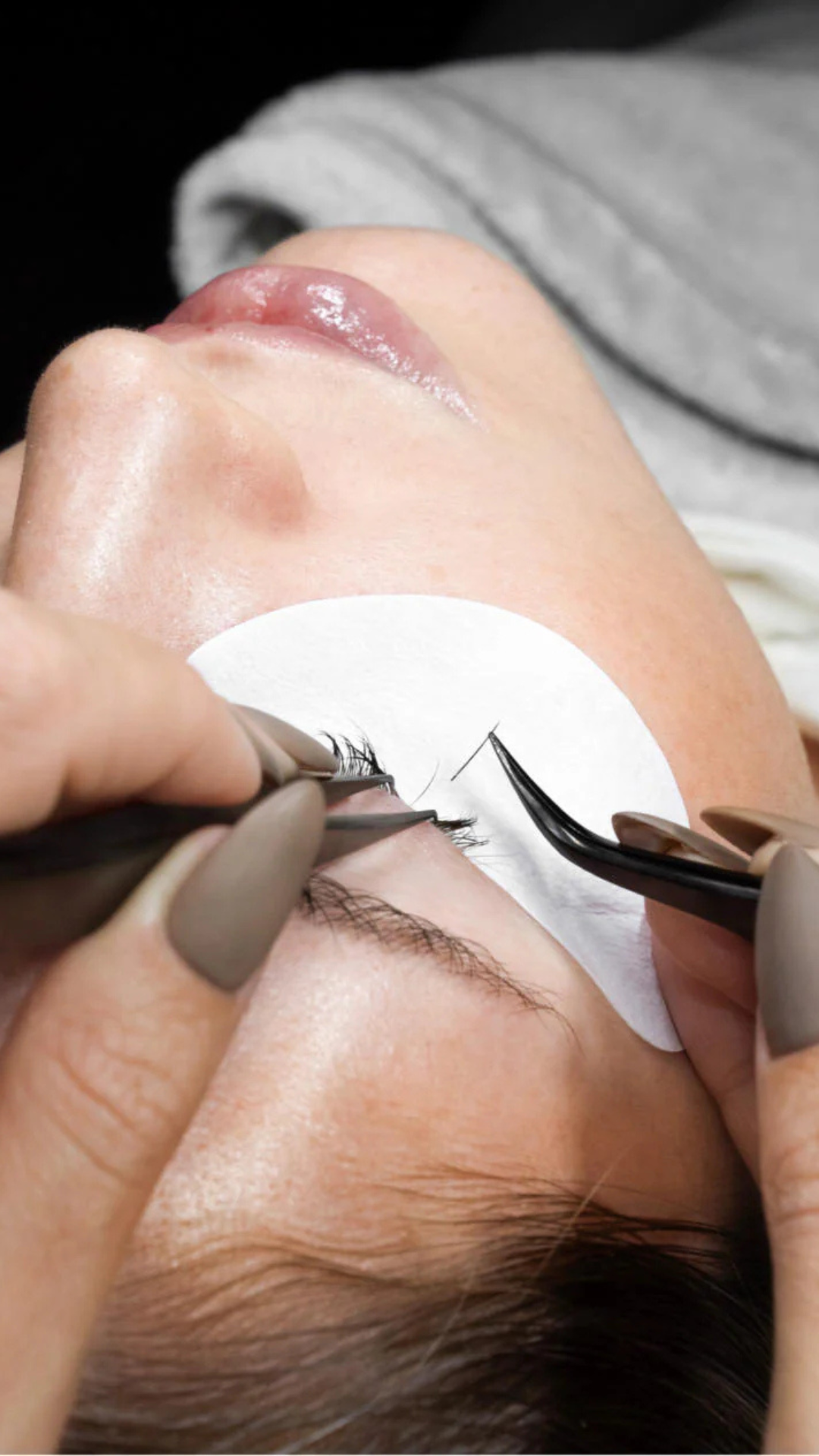 Close-up of a lash artist applying individual eyelash extensions with tweezers on a client’s eye, with an under-eye pad in place.