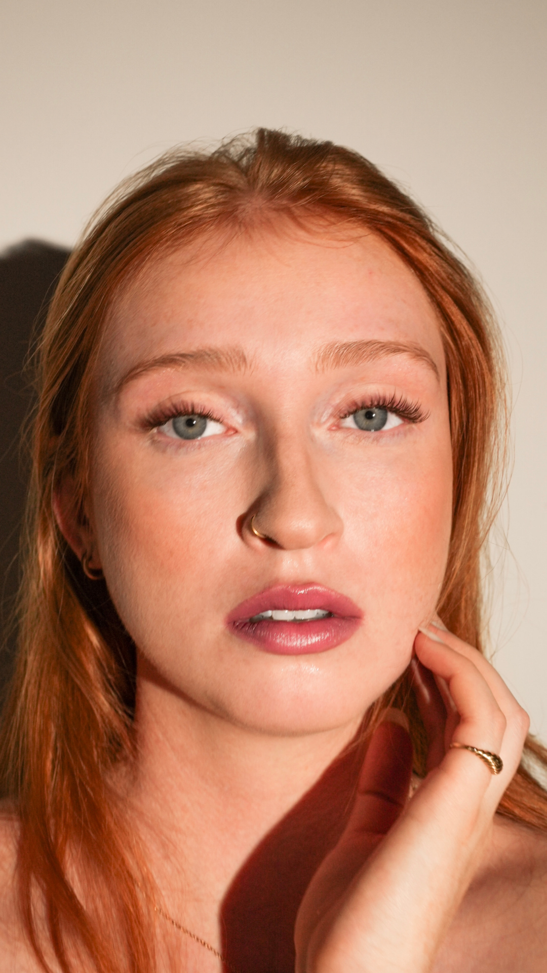 Portrait of a woman with long red hair, natural makeup, and glossy lips, looking directly at the camera with her hand resting near her face.