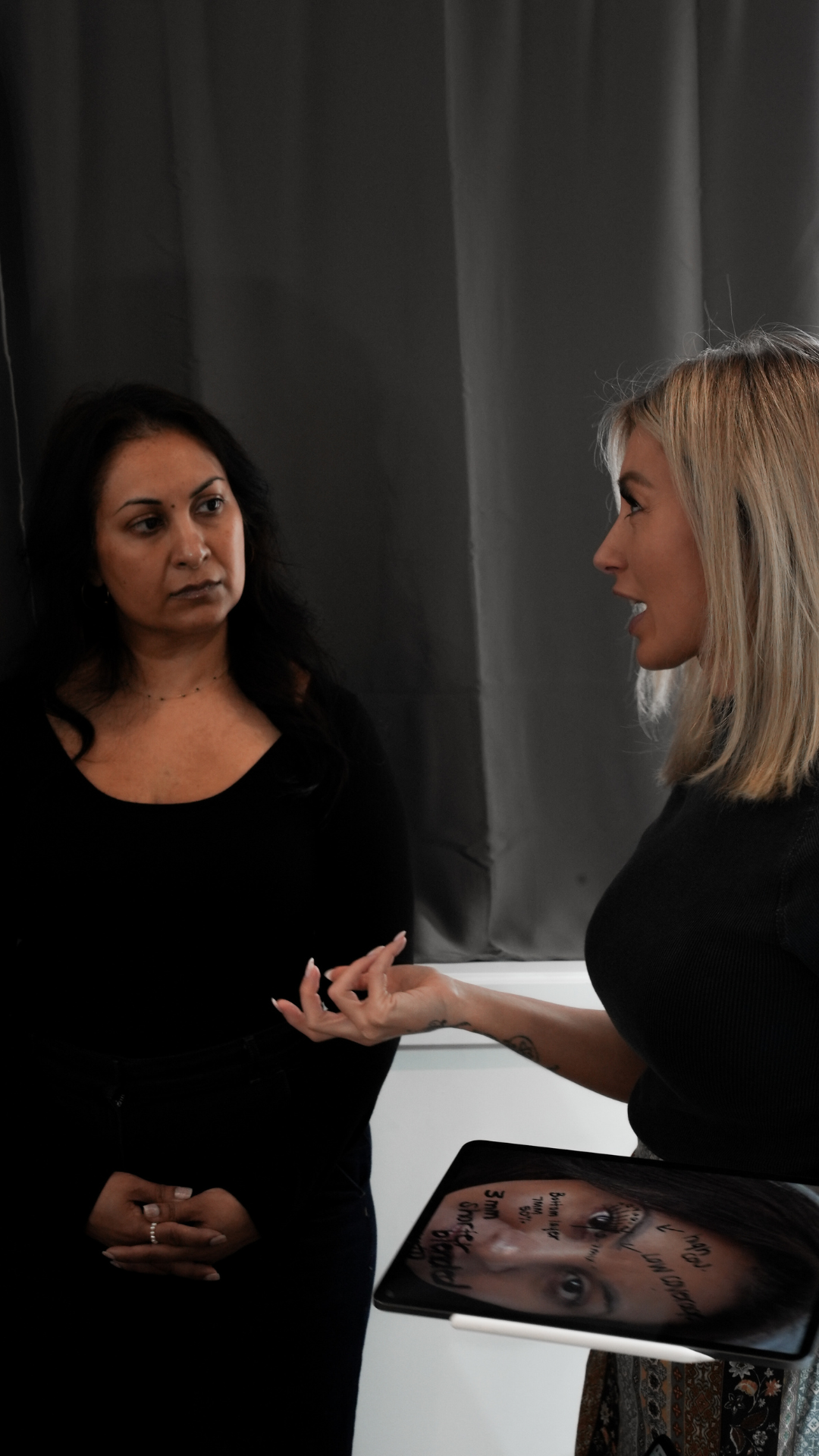 Two women in conversation indoors, one holding a tablet displaying a face diagram with lash mapping notes, suggesting a training or consultation setting.