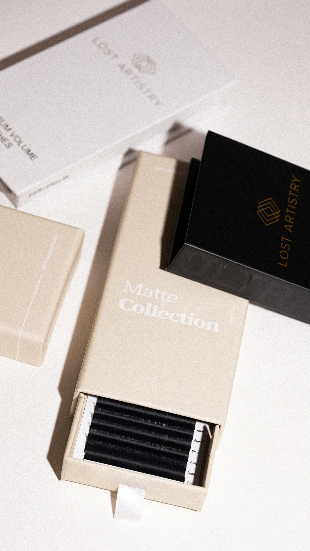 Lost Artistry Matte Collection lash tray in beige packaging, partially open to show black lash strips, with additional branded lash boxes in black and white nearby.