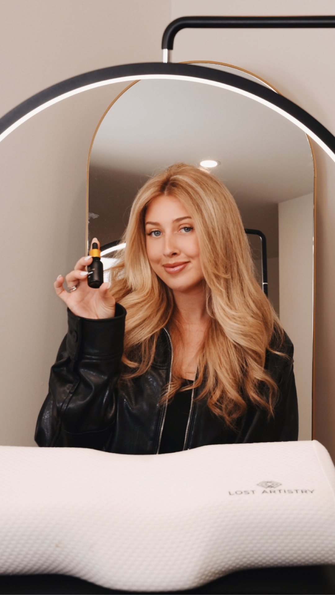 Smiling woman holding Lost Artistry’s Foreplay product in front of a mirror, with a Lost Artistry pillow in front of her