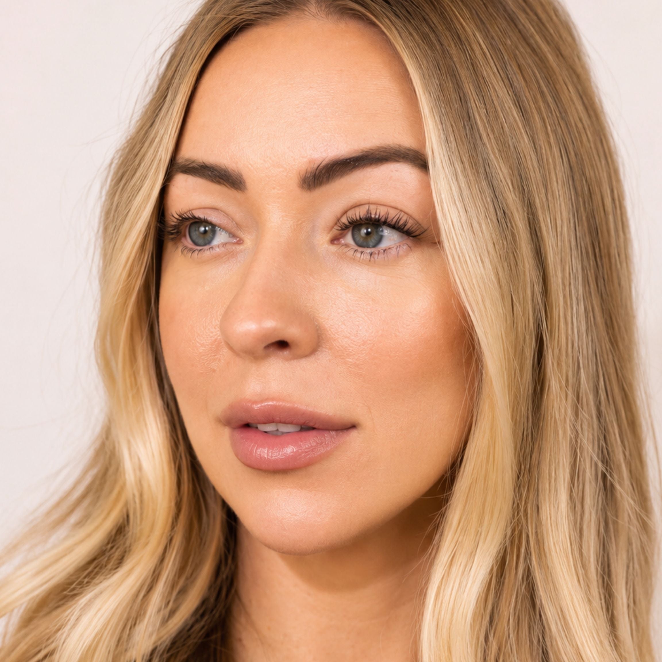 A close-up portrait of a woman with blonde hair, wearing a nude pink lip gloss and subtle makeup, set against a plain light background.