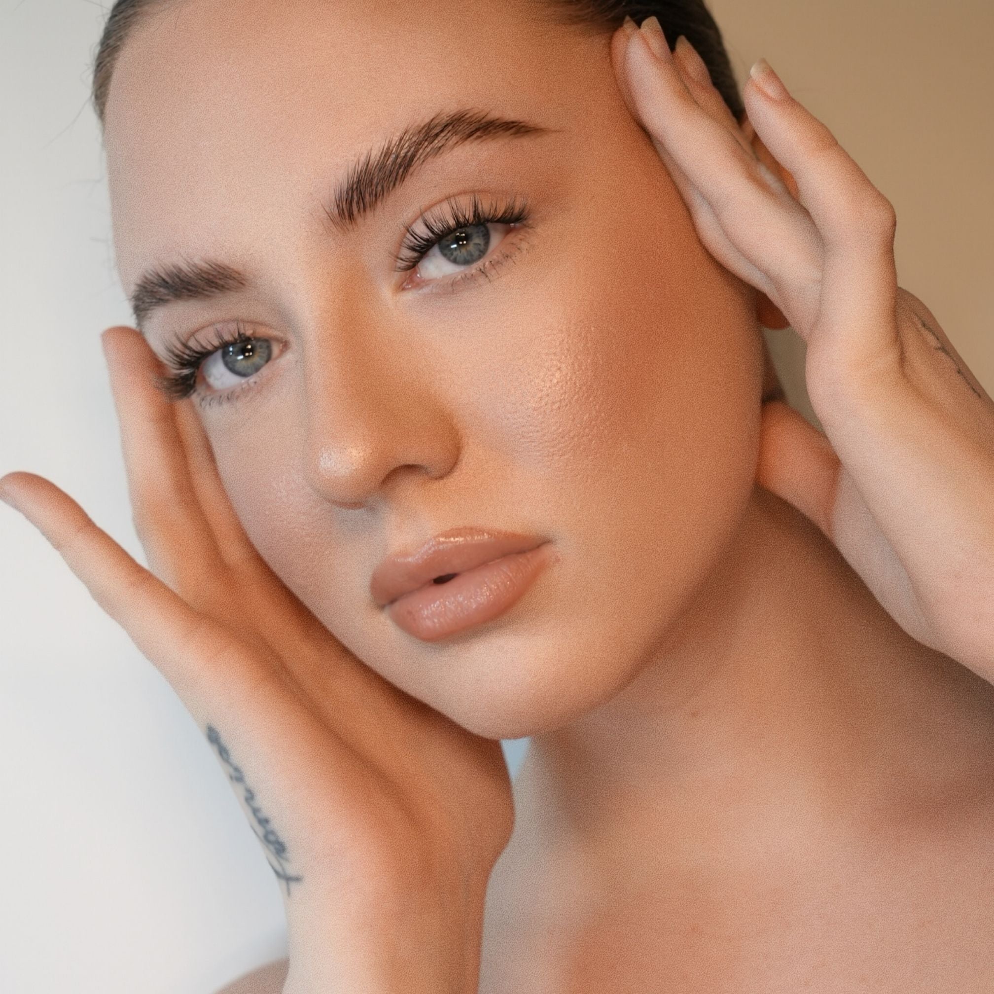 A close-up portrait of a woman with flawless, warm-toned makeup and defined eyebrows, holding her face with both hands against a soft beige background.