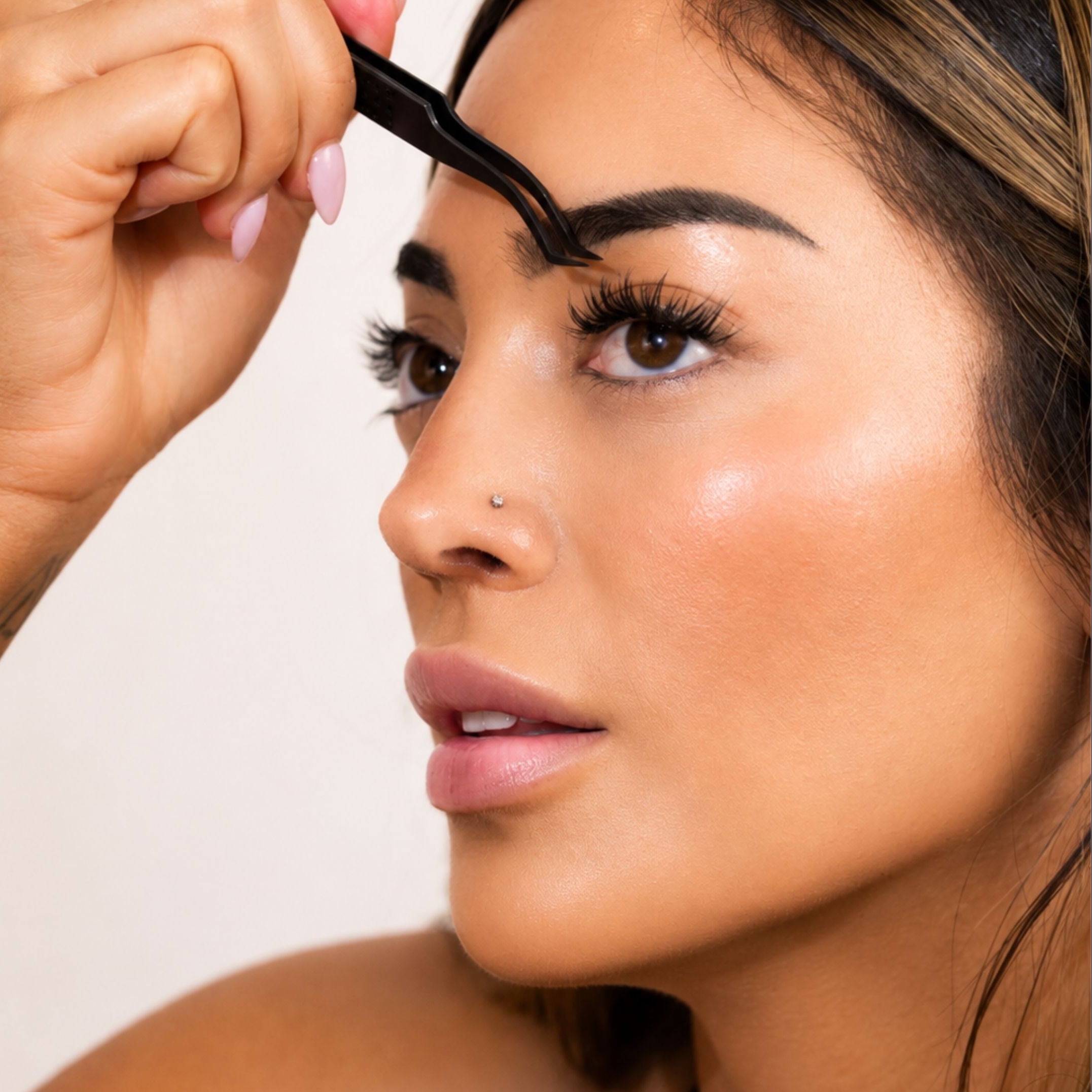 Close-up of a woman applying eyebrow makeup with a brow pencil, showing her face, hand, and hair against a plain light background.