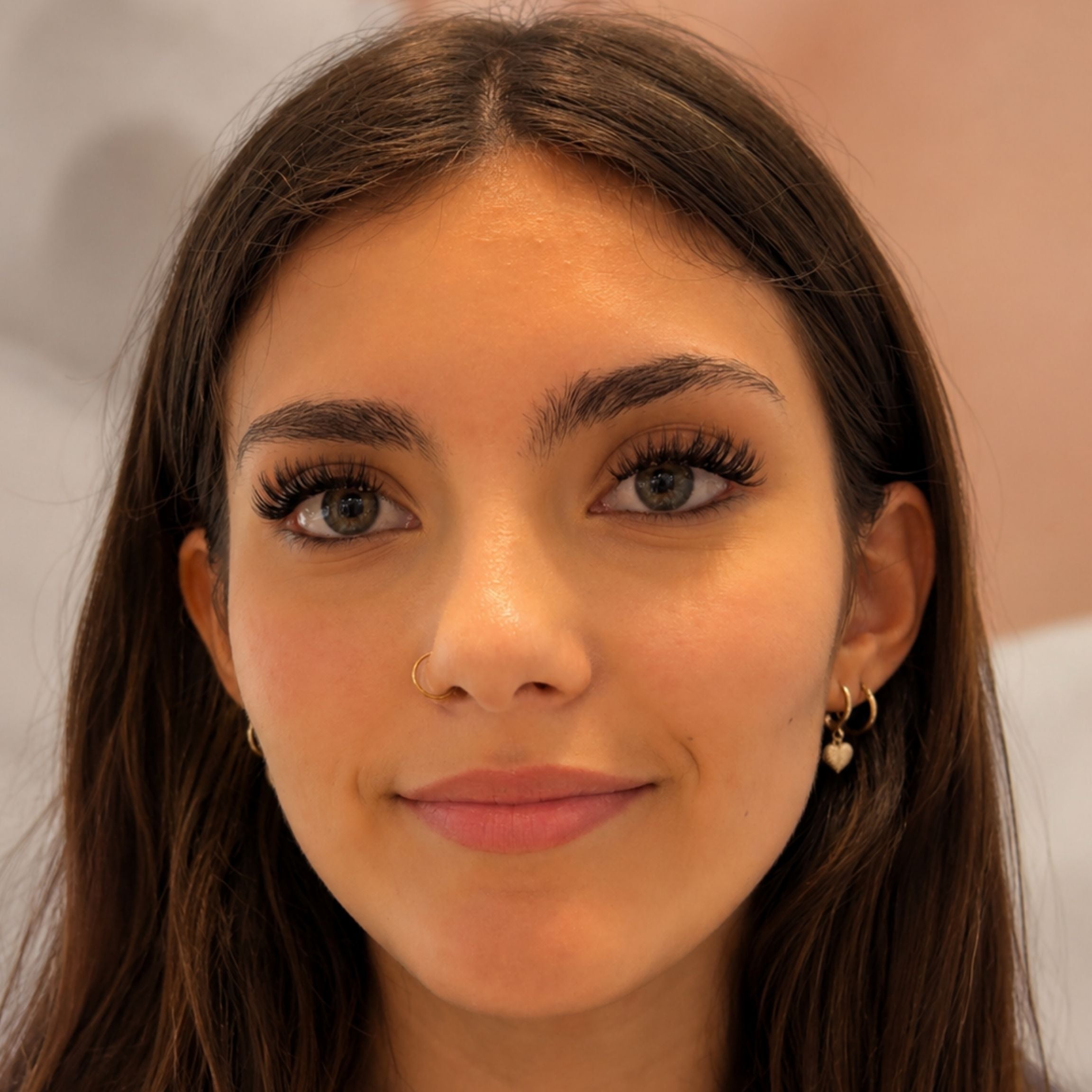 A close-up portrait of a woman with long dark brown hair, wearing small gold hoop earrings with white pearl details. She has long eyelashes, defined eyebrows, and is looking directly at the camera against a soft, out-of-focus background.