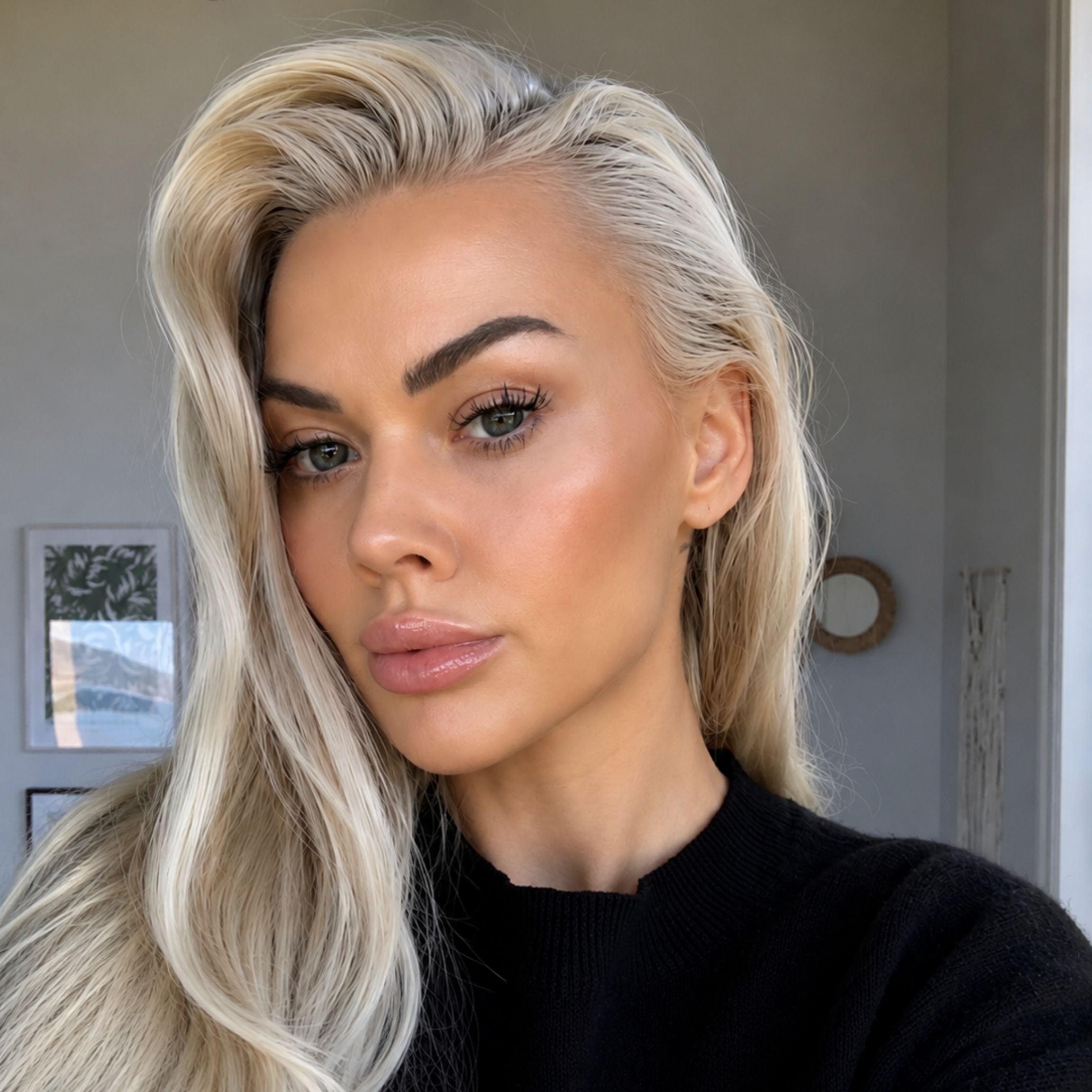 A close-up portrait of a woman with long, wavy platinum blonde hair, wearing a black knit top in an indoor setting.