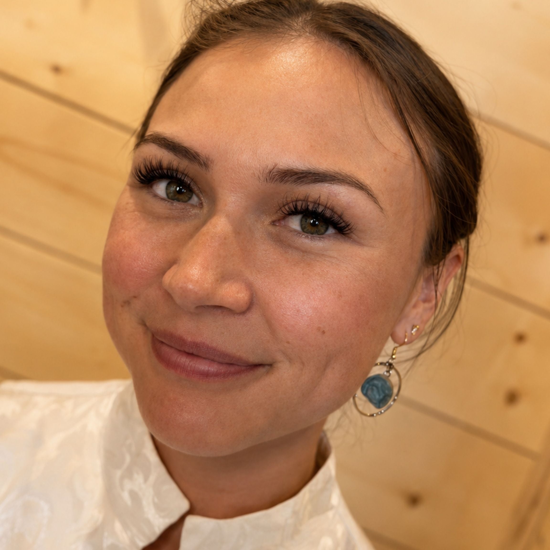 A close-up portrait of a woman with brown hair pulled back, wearing a white collared top and a dangling earring with a blue oval stone in a silver-toned setting, set against a light wood-paneled background.