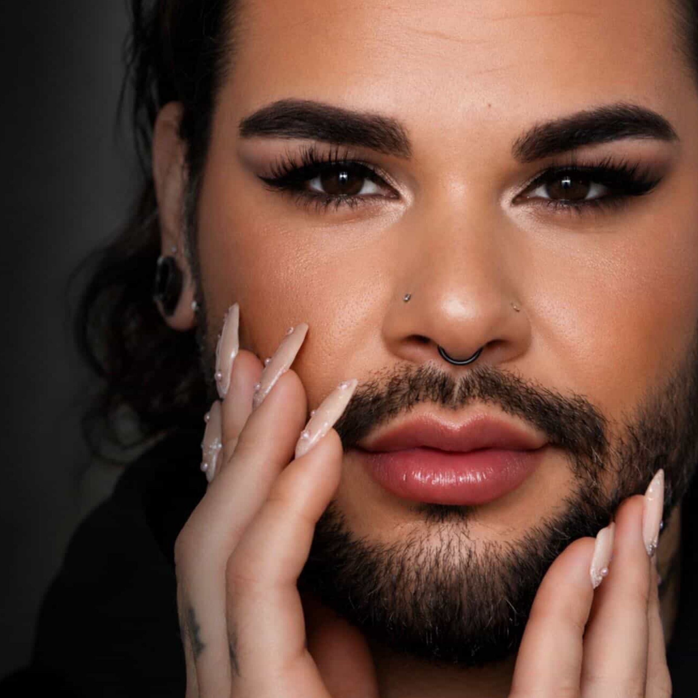 Close-up portrait of a person with dramatic eye makeup, a black nose ring, multiple facial piercings, styled beard, and long manicured nails touching their face against a dark studio background.