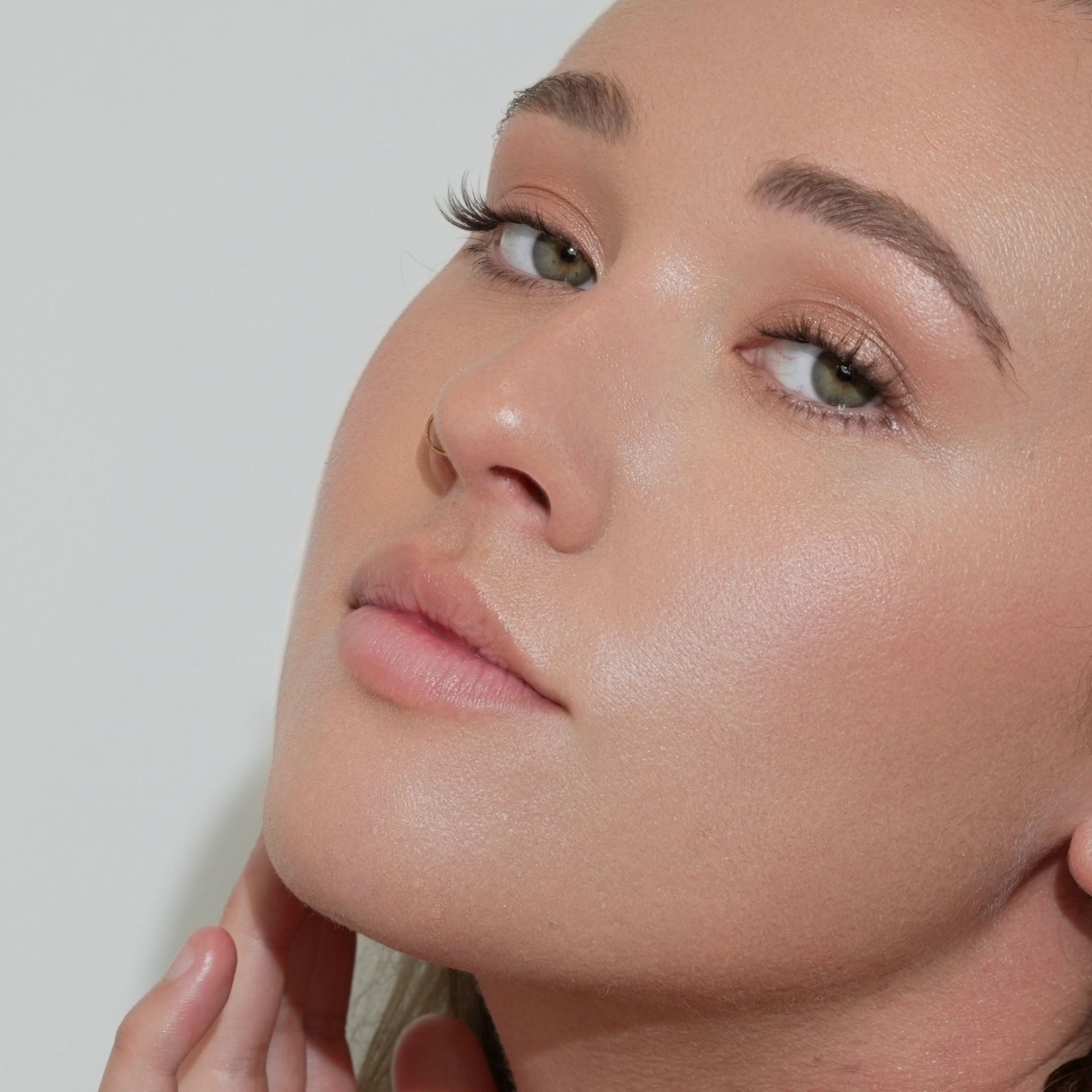 A close-up of a woman's face showing a high-shine, dewy finish on her skin, likely demonstrating a skincare or makeup product. She is looking upwards against a plain light background.