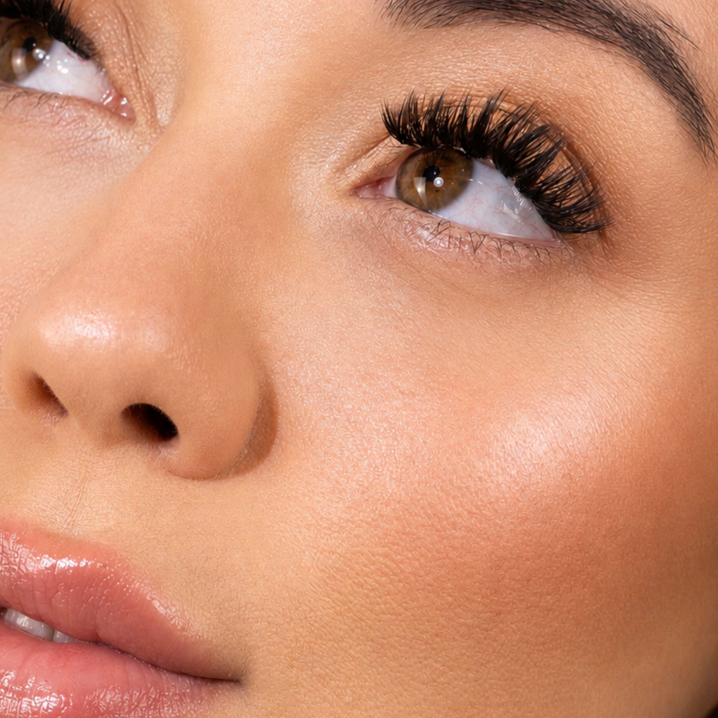 Extreme close-up of a woman's face highlighting long, voluminous black false eyelashes, smooth warm-toned skin, and glossy lips.