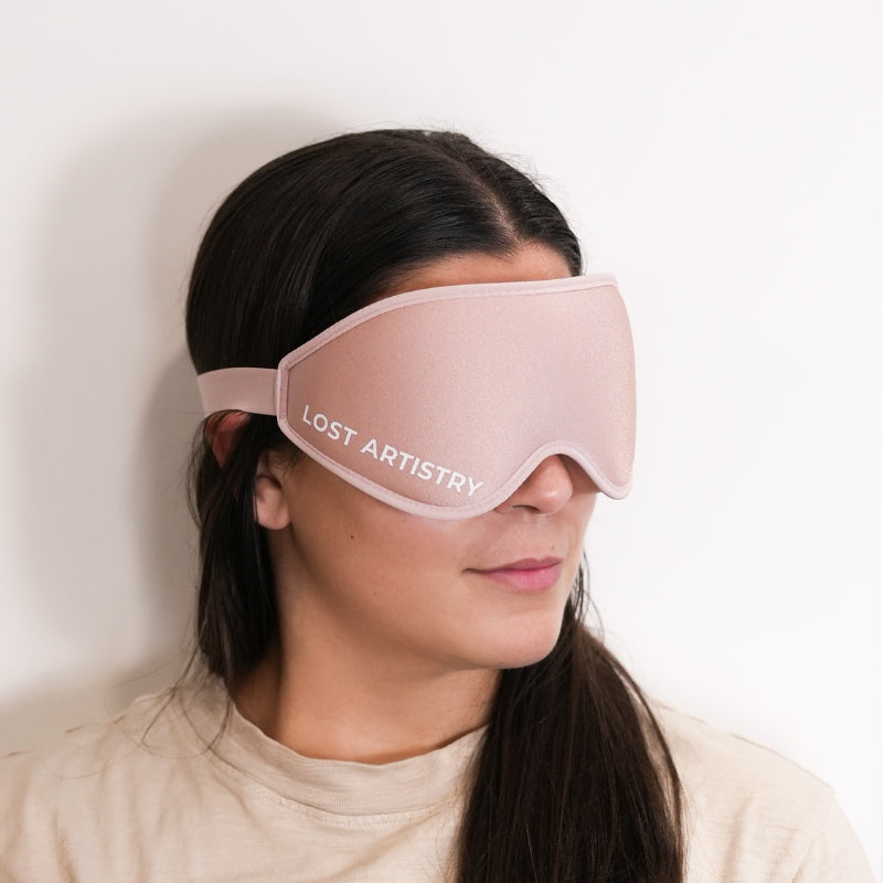 Person wearing a pink sleep mask with 'Lost Artistry' branding on a plain background